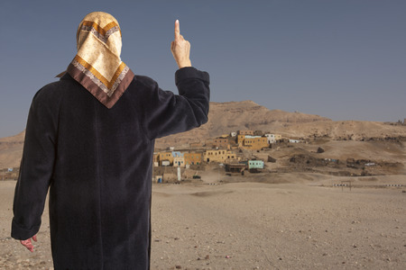 42985203 - muslim woman dressed in a black coat and scarf showing the raised pointer finger of her right hand in front of an arabic village in the desert.