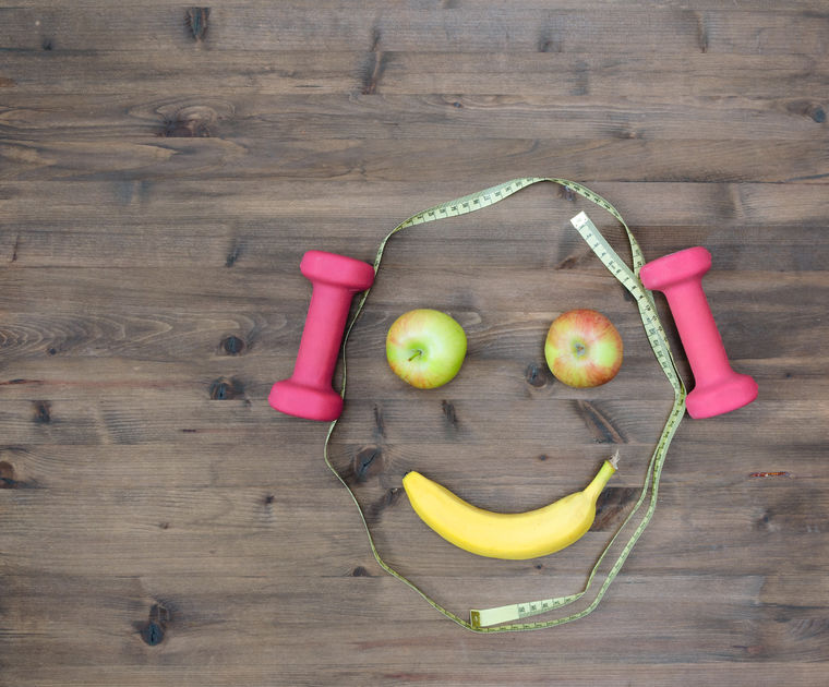 47751786 - healthy lifestyle concept. colored apples measuring tape dumbbells banana look like face on wooden table