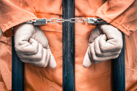 32219834 - handcuffed hands of a prisoner behind the bars of a prison with orange clothes - crispy desaturated dramatic filtered look