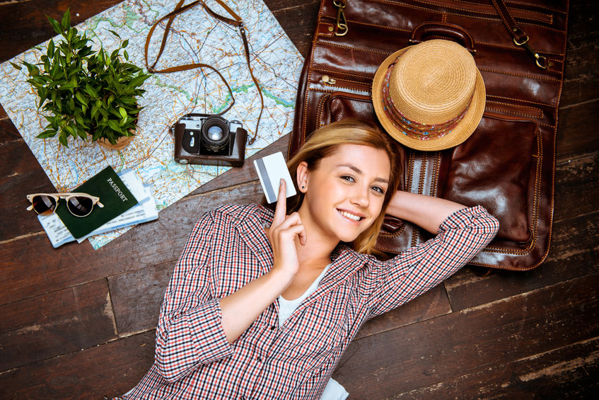 46697408 - top view photo of beautiful blonde girl lying on wooden floor. young woman smiling, holding credit card and looking at camera. passport, tickets, vintage camera, hat and map are on floor