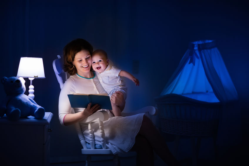 53287763 - mother and baby reading a book in dark bedroom. mom and child read books before bed time. family in the evening. kids room interior with night lamp and bassinet. parent holding infant next to crib.