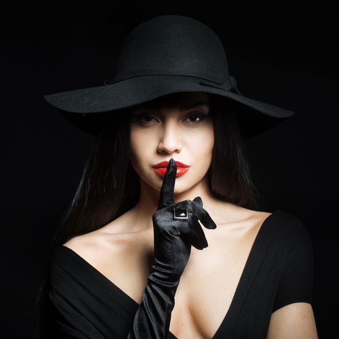 44816380 - woman in big black hat making a silence gesture, studio portrait, dark background