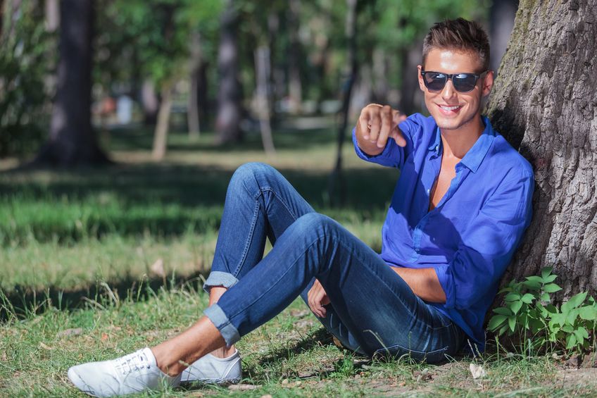 18600636 - young casual man sitting on the ground next to a tree and pointing at the camera while smiling