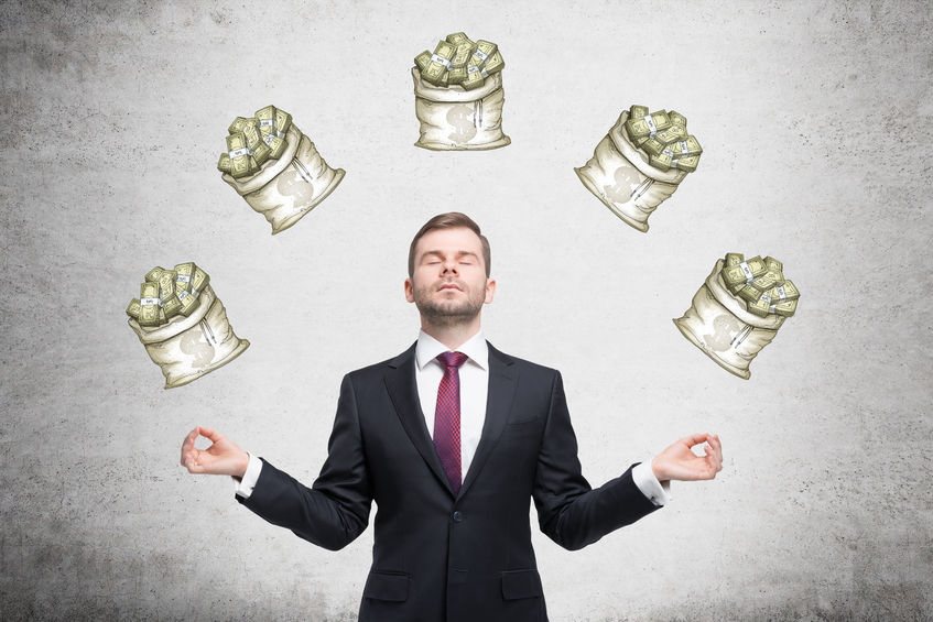 52395260 - a young businessman standing in the posture of meditation with eyes closed at a concrete wall with five sacks of dollars on it. front view. concept of dreaming about money.