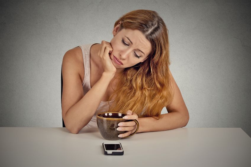 36468116 - sad depressed young woman sitting at table drinking coffee looking at her mobile phone waiting for a call text message isolated on grey wall background.