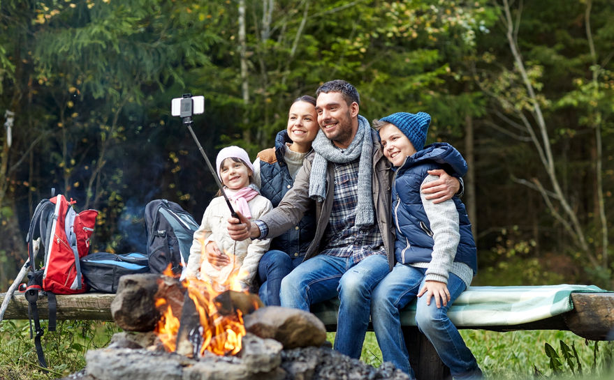 49276540 - camping, travel, tourism, hike and people concept - happy family sitting on bench and taking picture with smartphone on selfie stick at campfire in woods