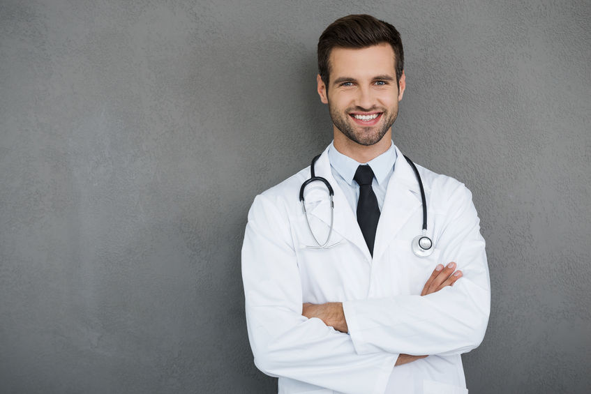 44203305 - you can absolutely trust me. confident young doctor in white uniform looking at camera and keeping arms crossed while standing against grey background