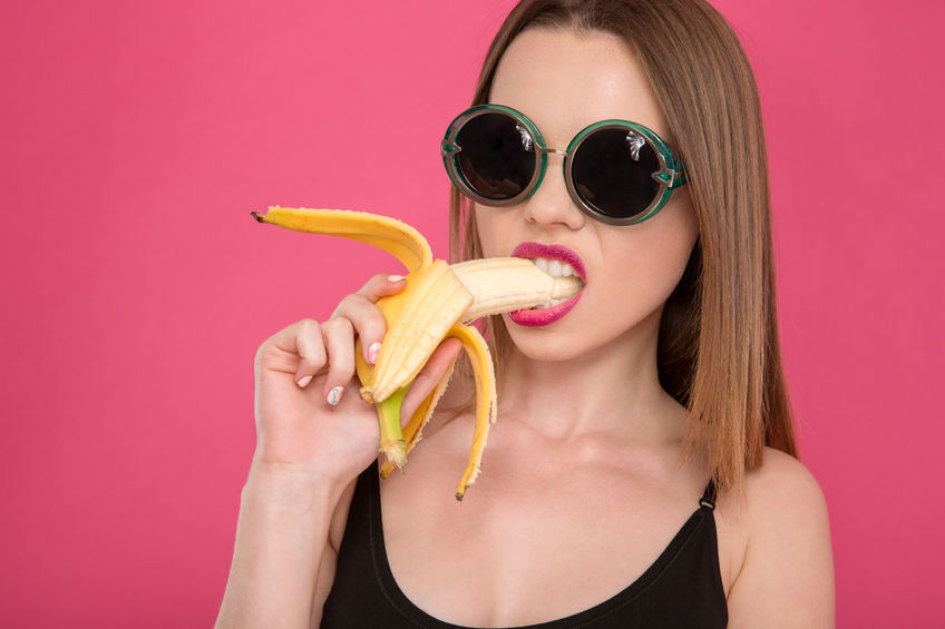 48376499 - closeup of young seductive attractive sensual model in black sunglasses eating banana on pink background